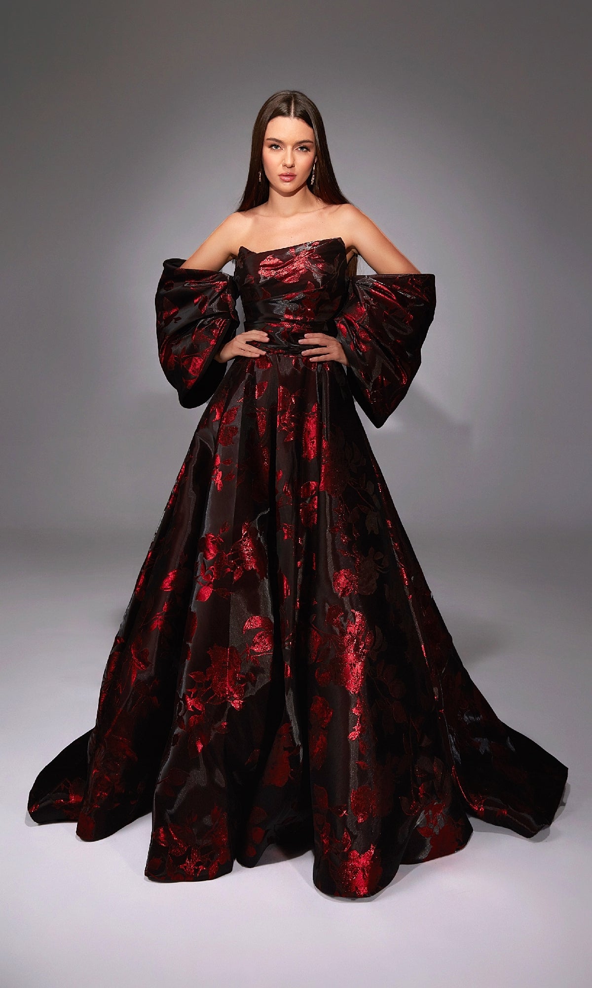 A woman poses in a studio wearing the Alyce 27862, a long formal off-the-shoulder floral jacquard gown with red patterns and dramatic puffed sleeves—an ideal choice for any formal ball.